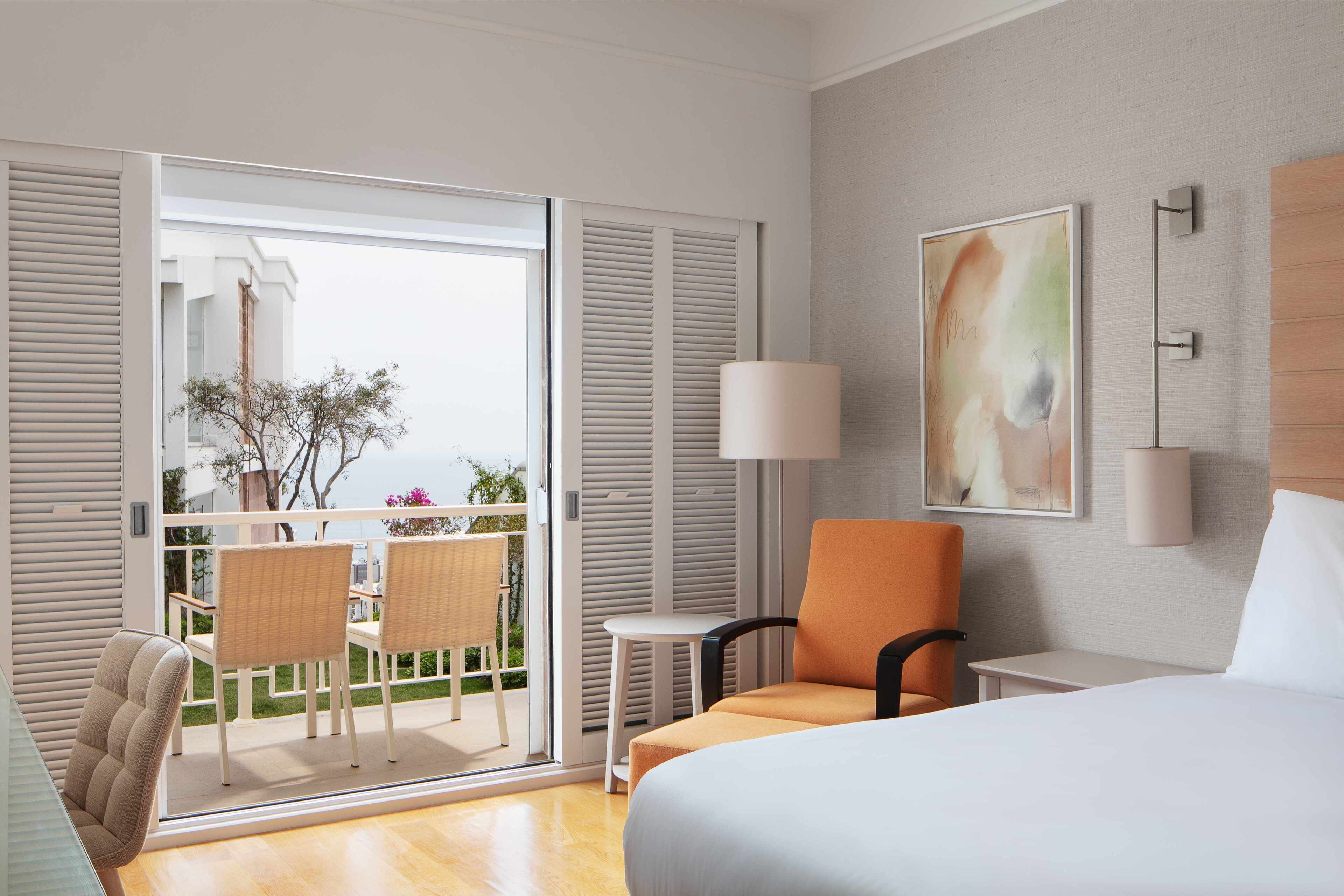 Hotelkamer met een bed, een stoel, een klein tafeltje, een balkon met stoelen en uitzicht op de omgeving.