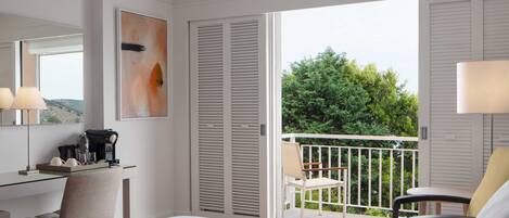 A hotel room with a bed, a desk with a coffee maker, a chair, a balcony with a view of trees, and a painting on the wall.