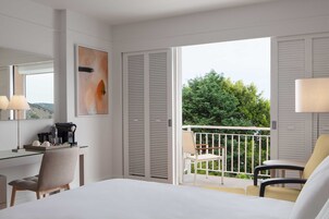 Habitación de hotel con cama, escritorio con cafetera, silla, balcón con vista a árboles y un cuadro en la pared.