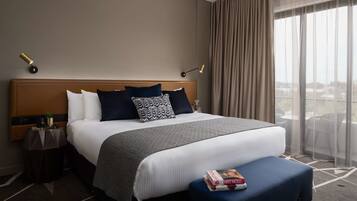 A hotel room with a large bed, a bedside table with a lamp, a window with curtains, and a view of the city.