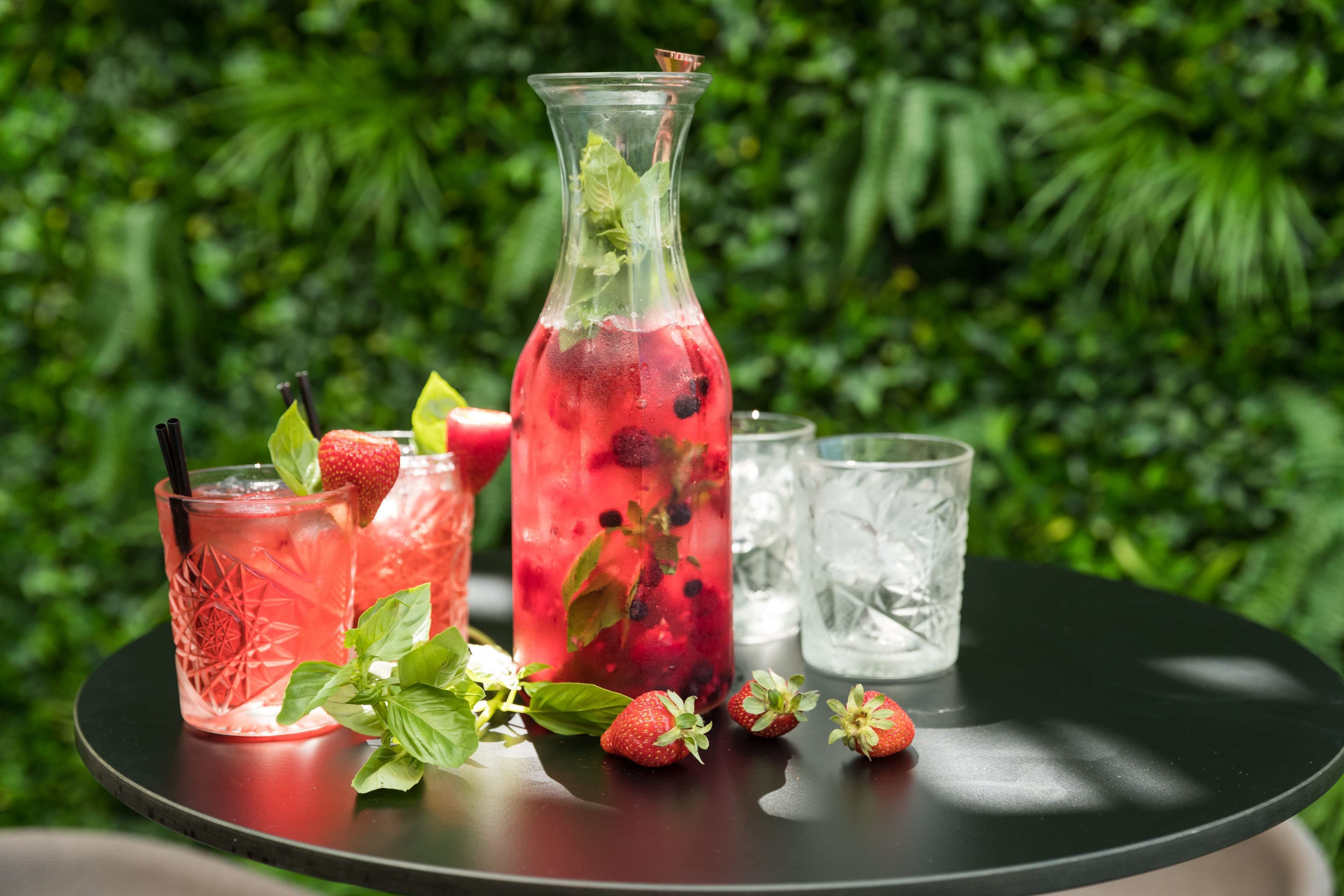 Una mesa con una jarra y vasos de una bebida roja, fresas y hojas de menta.