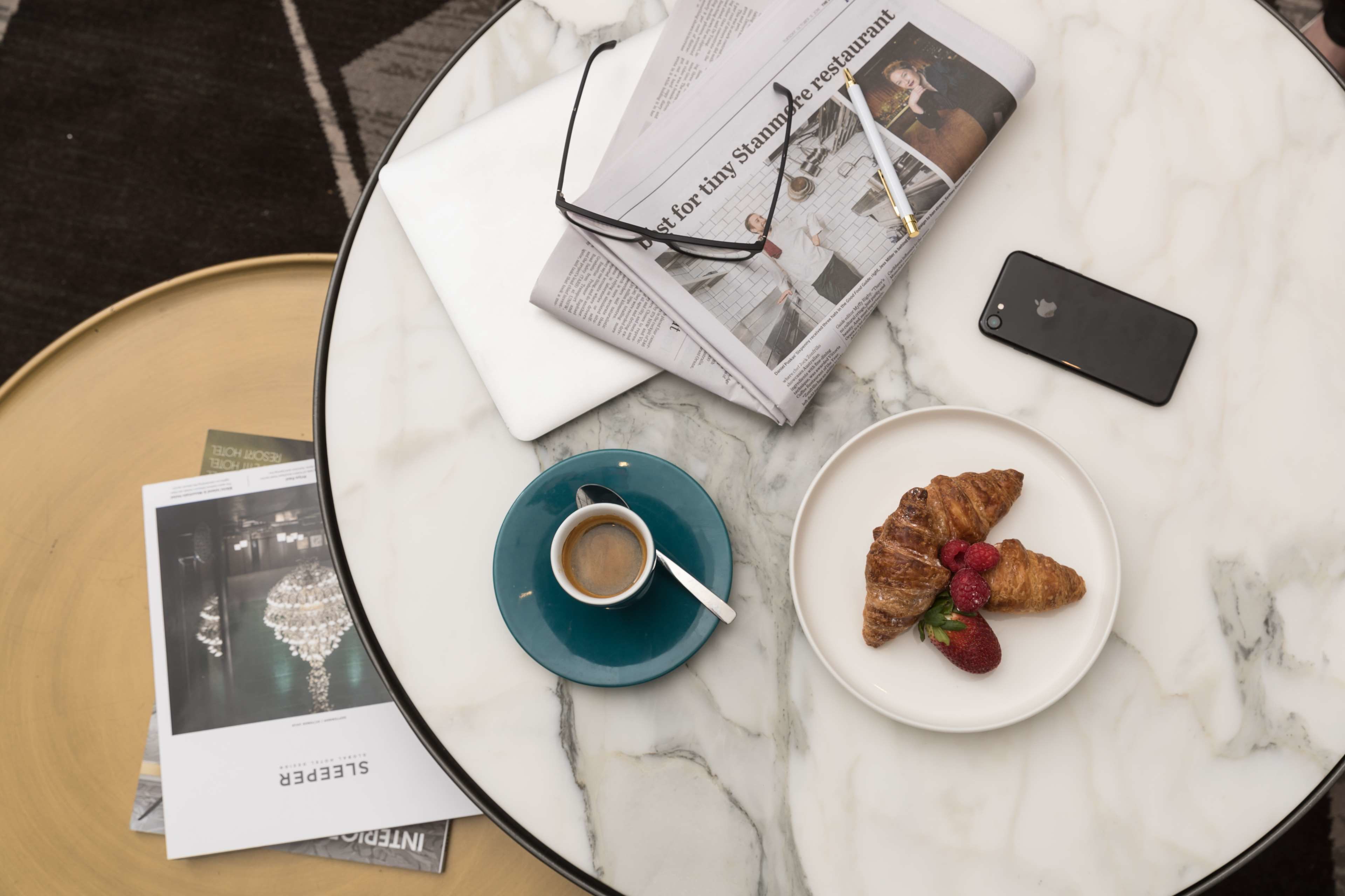 A round table with a laptop, a smartphone, a plate with a croissant and strawberries, a cup of coffee, a pair of glasses, and some brochures.