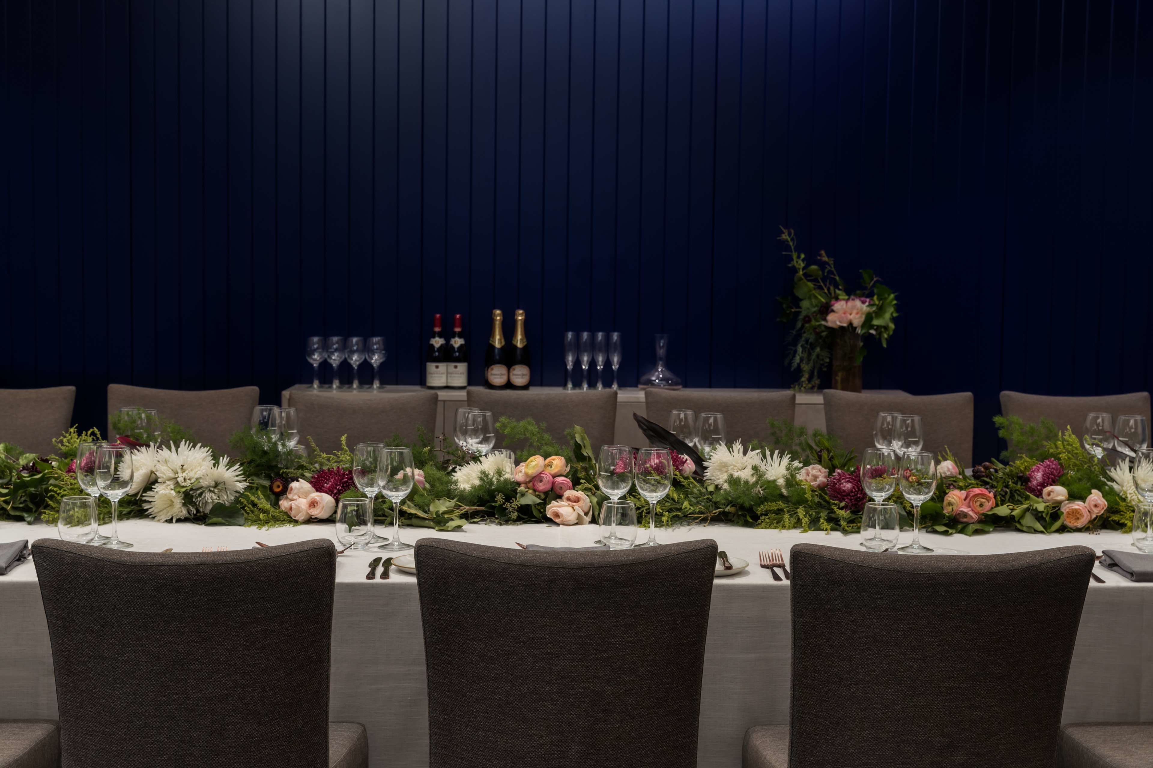 Una mesa de comedor formal dispuesta con múltiples copas de vino, botellas y centros de mesa florales.