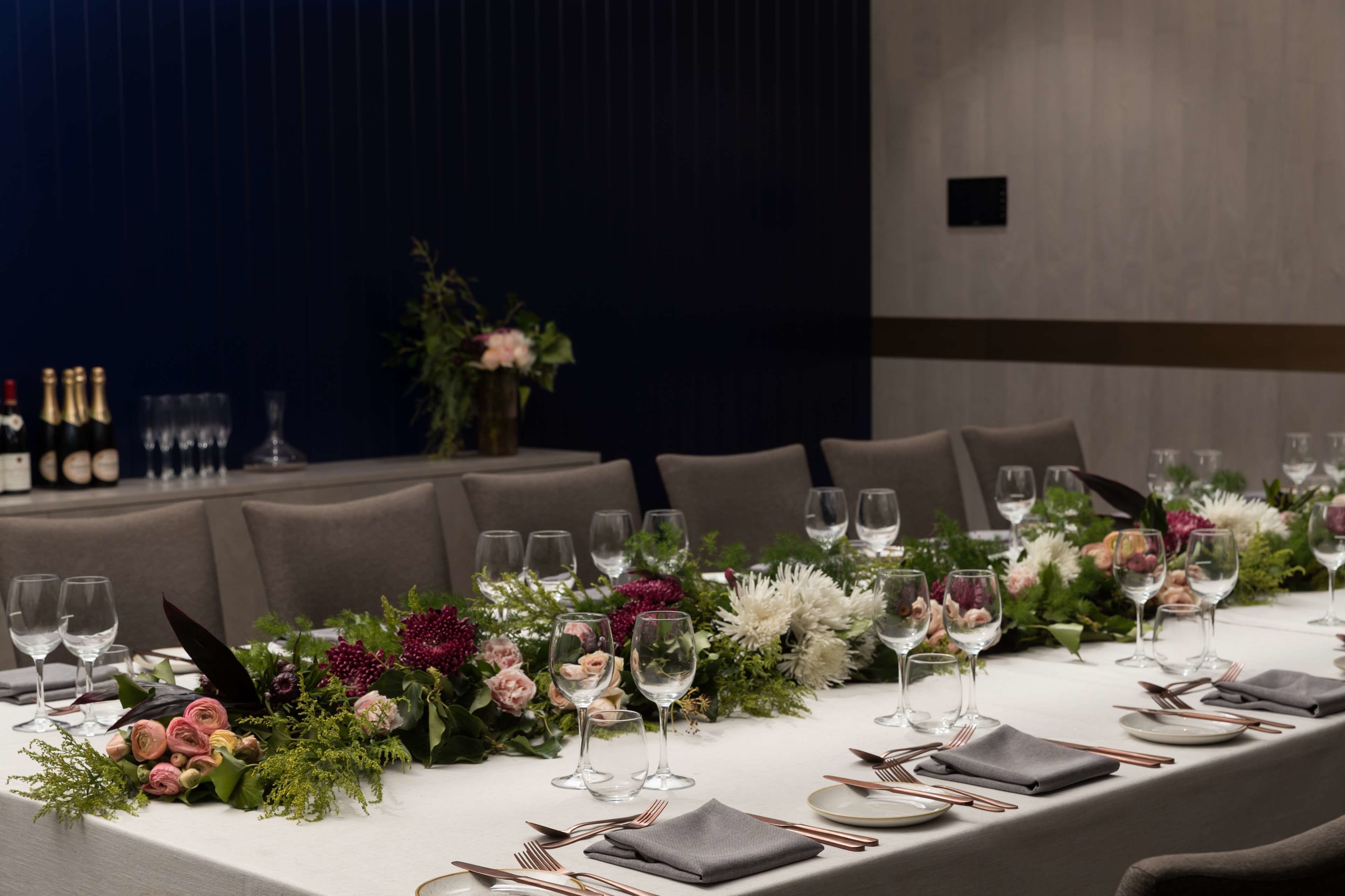 A long dining table set with multiple wine glasses, plates, and cutlery, surrounded by floral centerpieces.