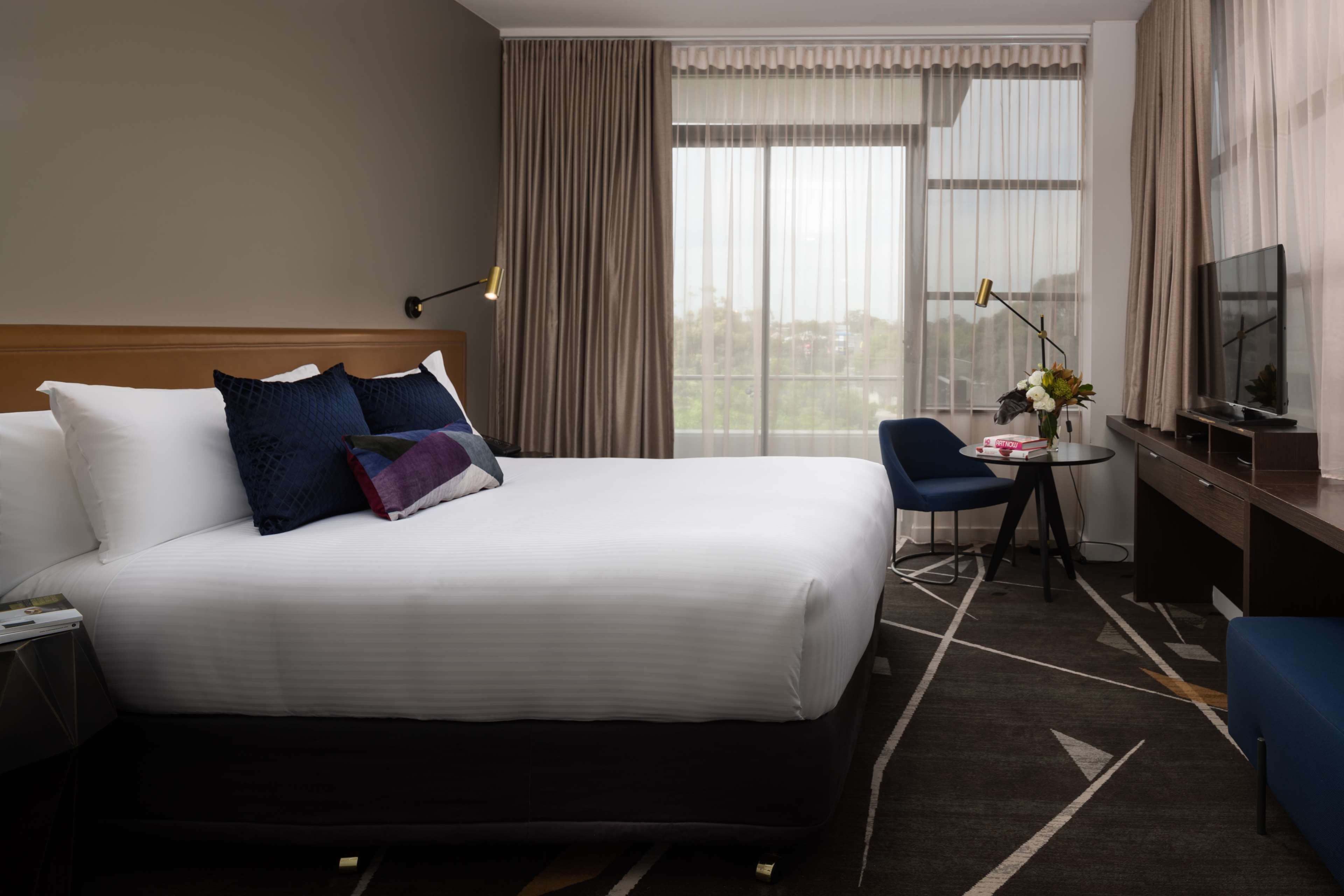 Habitación de hotel con una cama grande, un escritorio con silla, un televisor y una ventana con cortinas.
