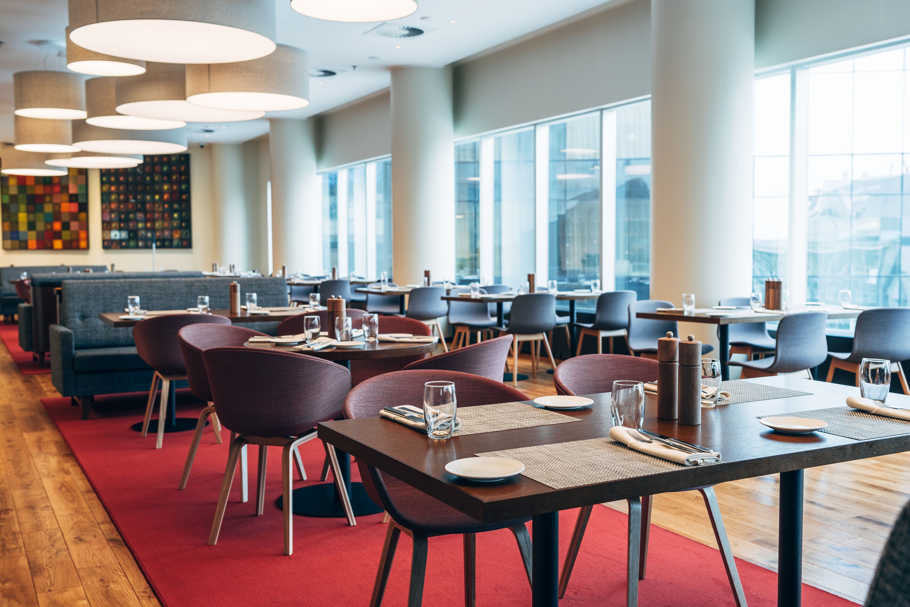 A modern restaurant with multiple dining tables and chairs, large windows, and a red carpet.
