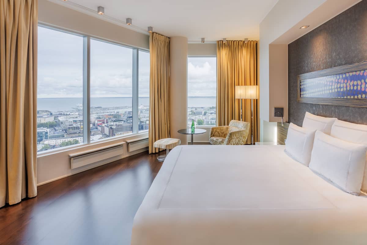 A hotel room with a large bed, a small round table, a chair, and a view of the city through large windows.