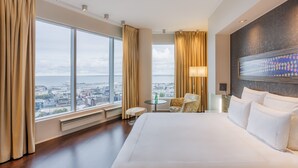 A hotel room with a large bed, a small round table, a chair, and a view of the city through large windows.