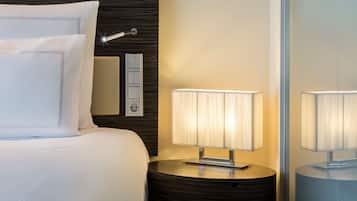 A neatly made bed with white linens and pillows, a bedside table with a lamp, and a wall-mounted control panel.