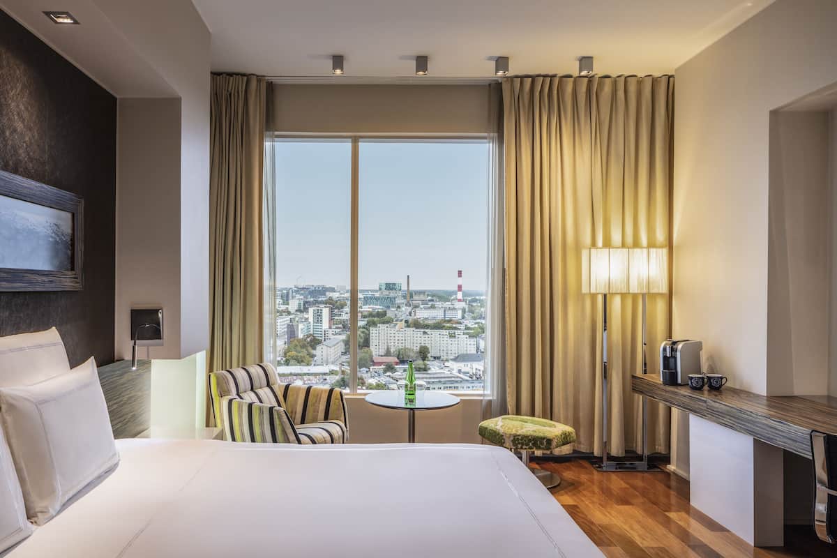 A hotel room with a large bed, a seating area with a chair and ottoman, a small round table with a bottle, a lamp, and a coffee maker on a wooden side table.