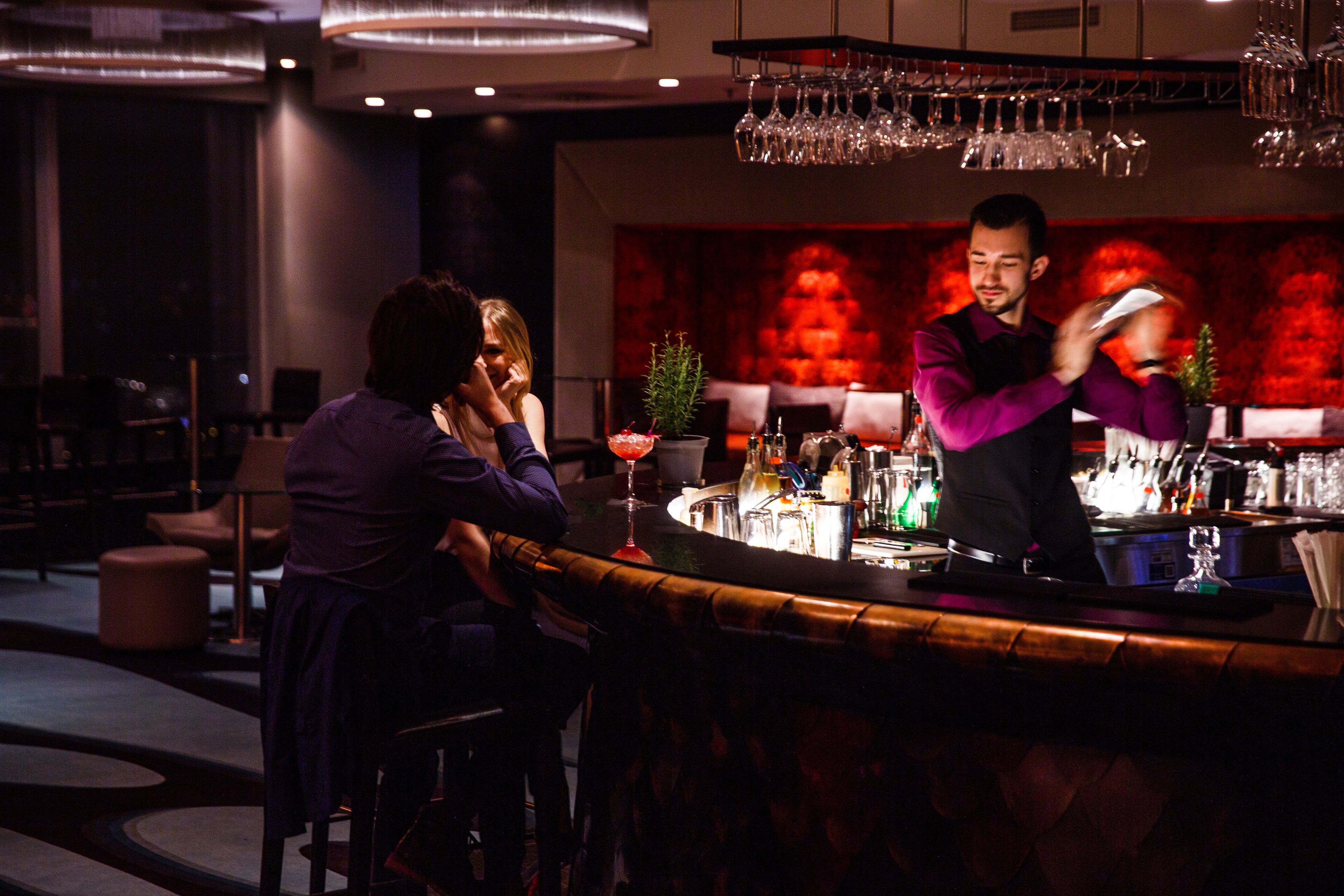 A bar area with a bartender preparing drinks, a bar counter, and a seating area with chairs and a table.