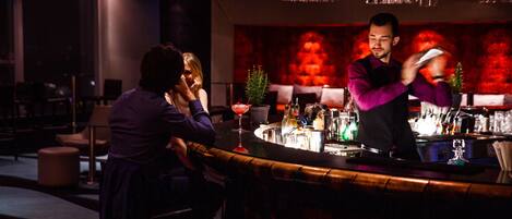 A bar area with a bartender preparing drinks, a bar counter, and a seating area with chairs and a table.