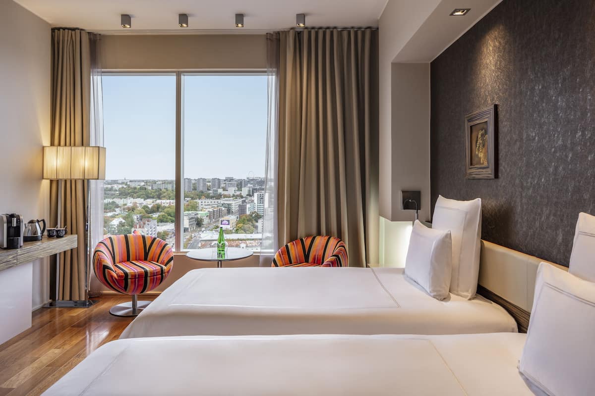A hotel room with two beds, a view of the city, a desk with a lamp, and a chair.
