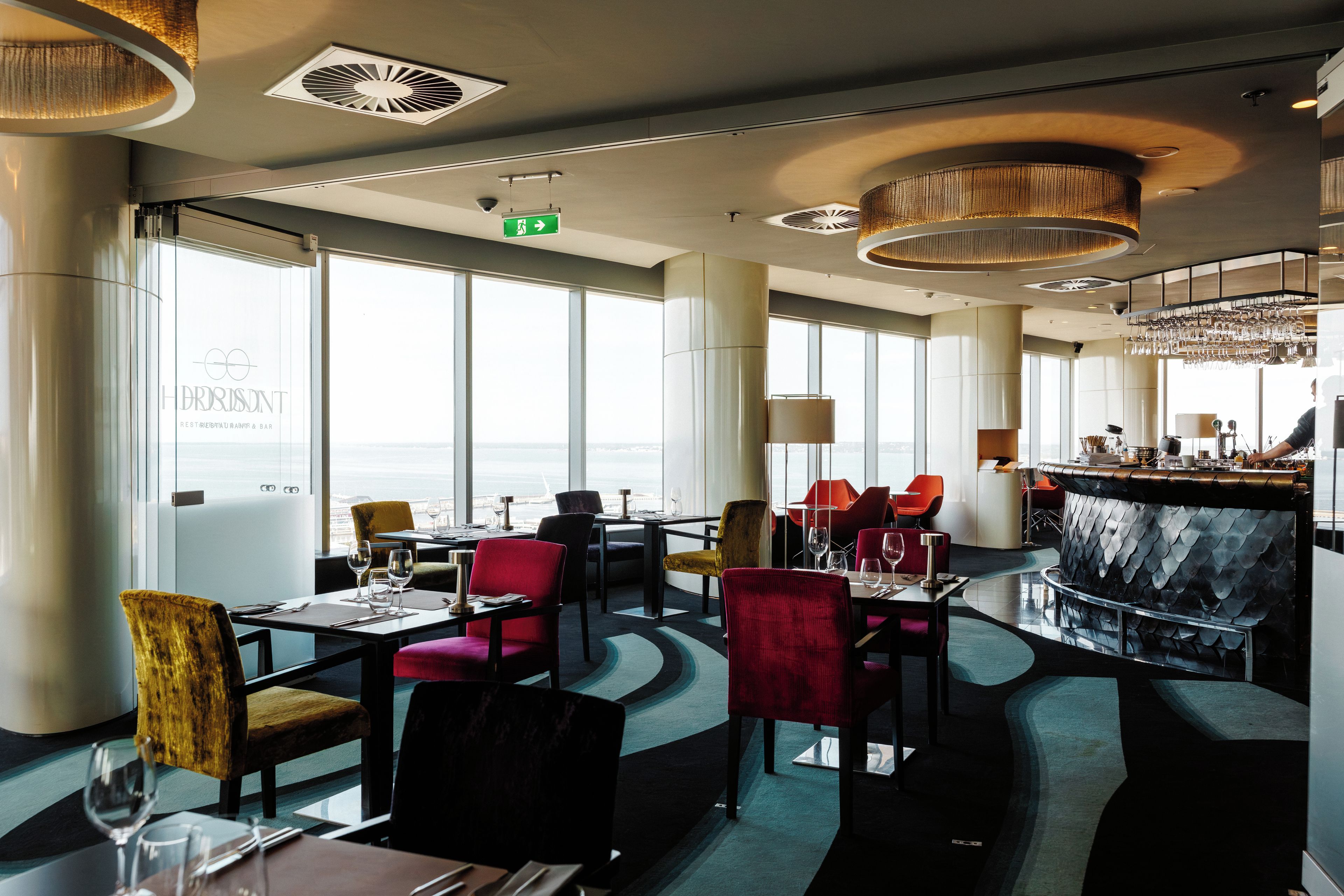 A modern restaurant interior with high ceilings, large windows, and a bar area.