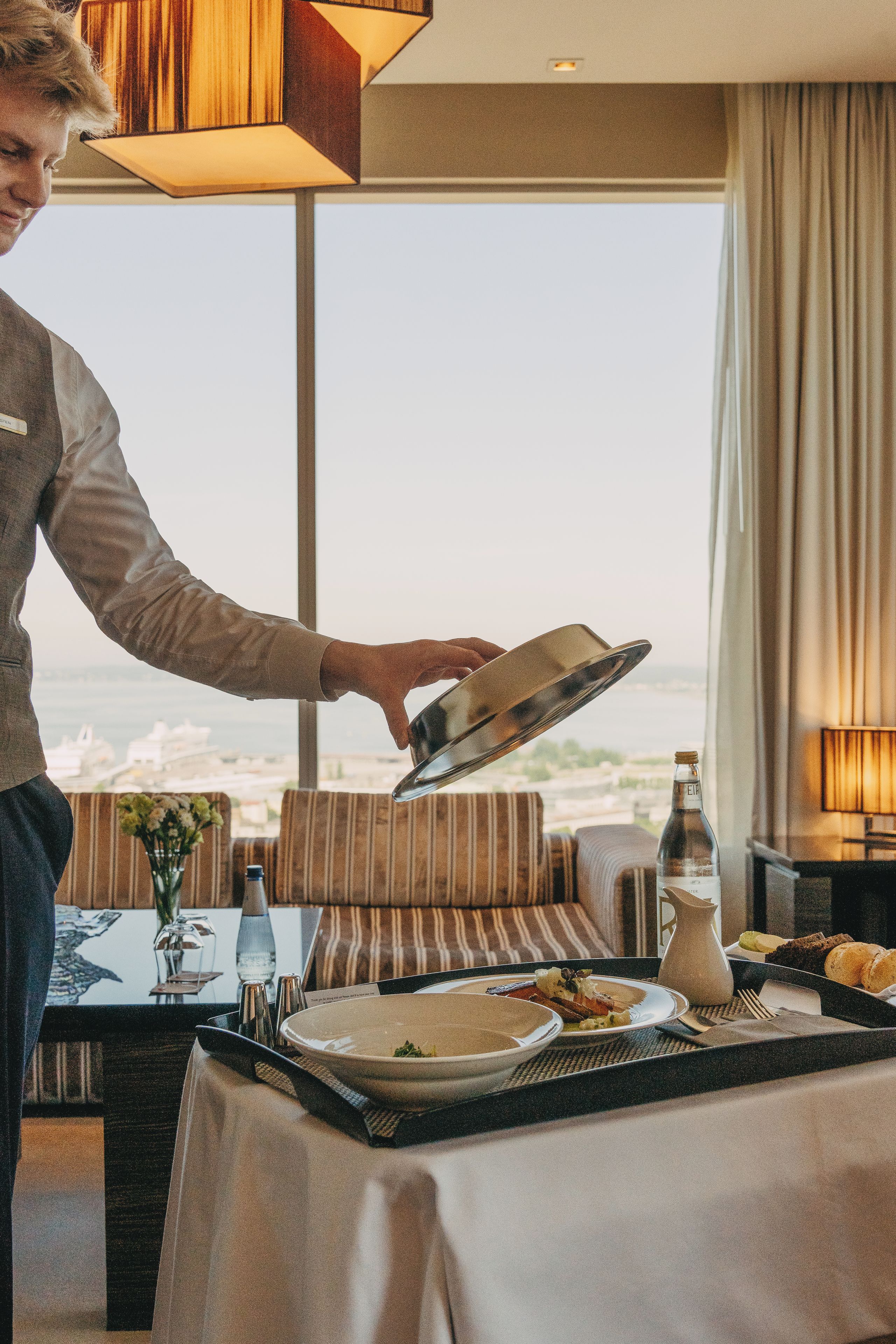 A hotel room with a dining table set for a meal, a view of the cityscape, and a large window with curtains.