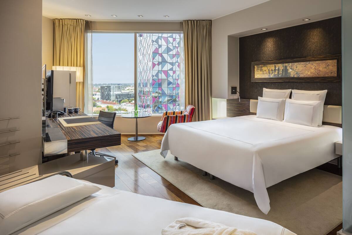 A hotel room with a large bed, a desk with a computer, a chair, and a view of the city through a window with a geometric pattern.
