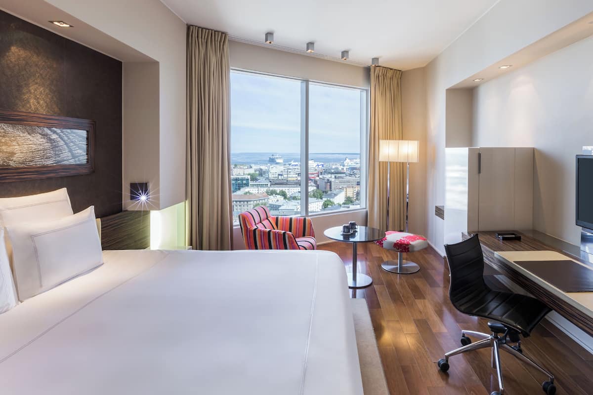 A hotel room with a large bed, a TV, a desk, a chair, a lamp, and a view of the city.
