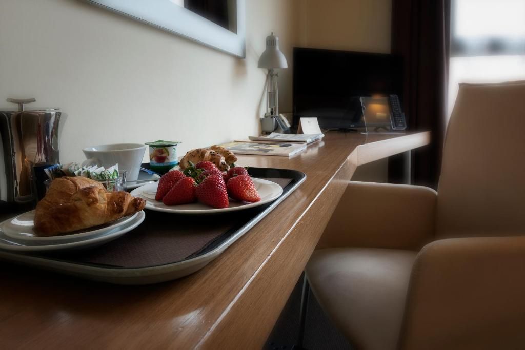 A hotel room with a dining table set with croissants and strawberries, a lamp, and a television.