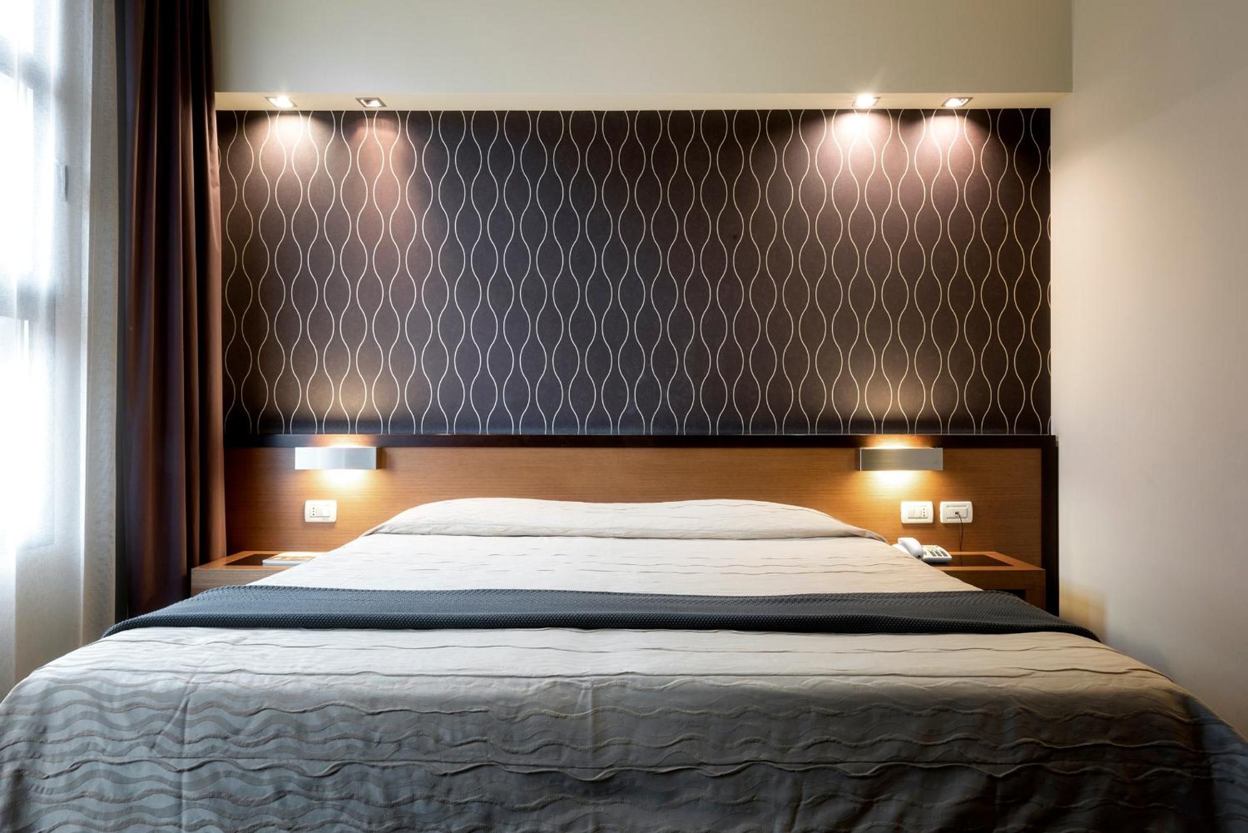 A hotel room with a bed, two bedside tables, a headboard with a patterned design, and wall-mounted lights.