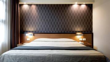 A hotel room with a bed, two bedside tables, a headboard with a patterned design, and wall-mounted lights.