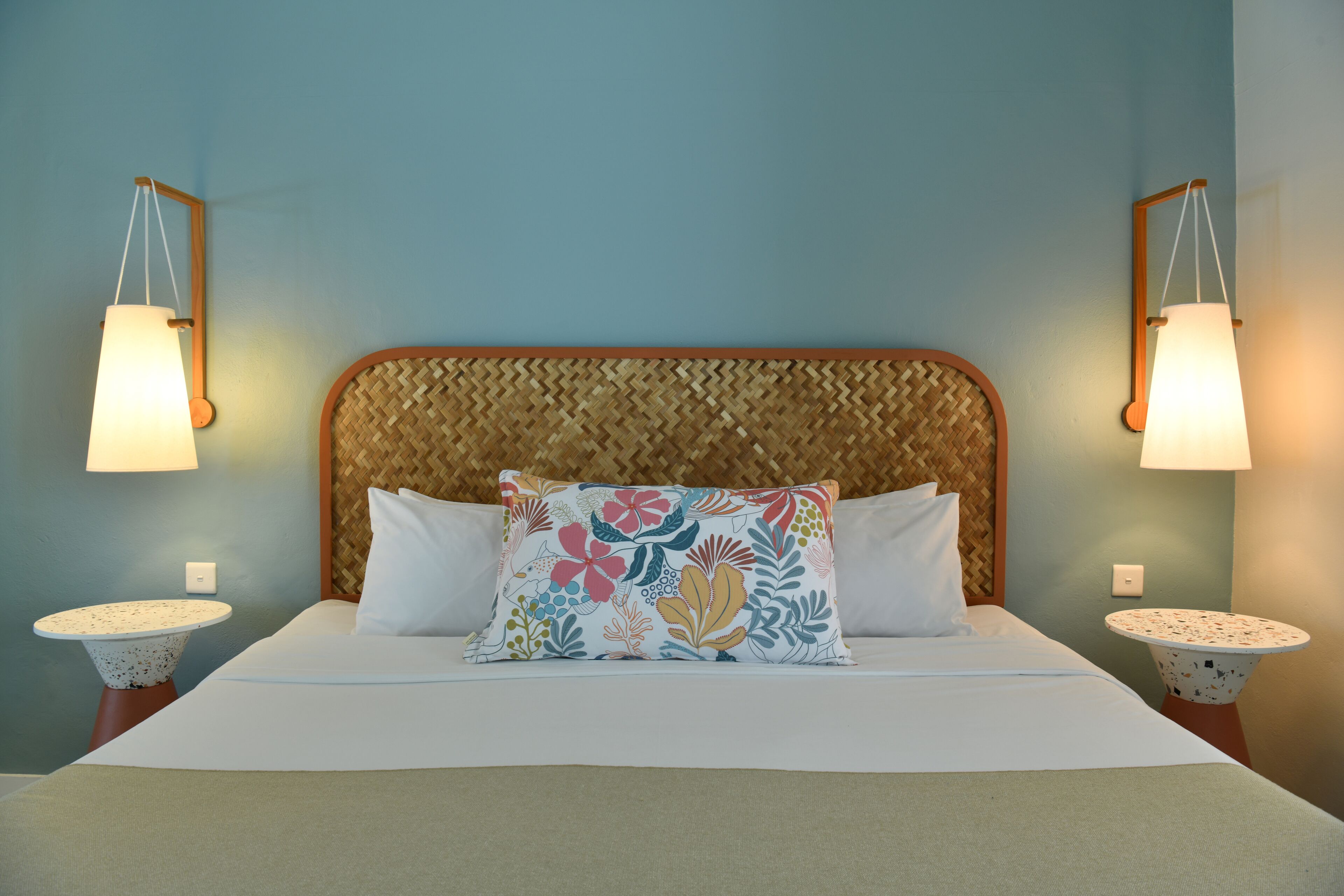 A neatly made bed with a floral pillow, two bedside tables with lamps, and a woven headboard.
