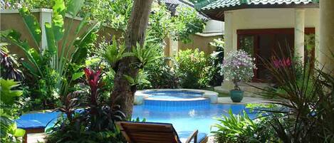 Zona de piscina con una silla de madera, rodeada de exuberante vegetación y una estructura de techo con tejas.
