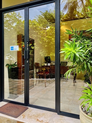 Entrada de hotel con puerta de vidrio, un cartel que indica que el hall está abierto y vistas del área de estar exterior con plantas y suelo de baldosas.