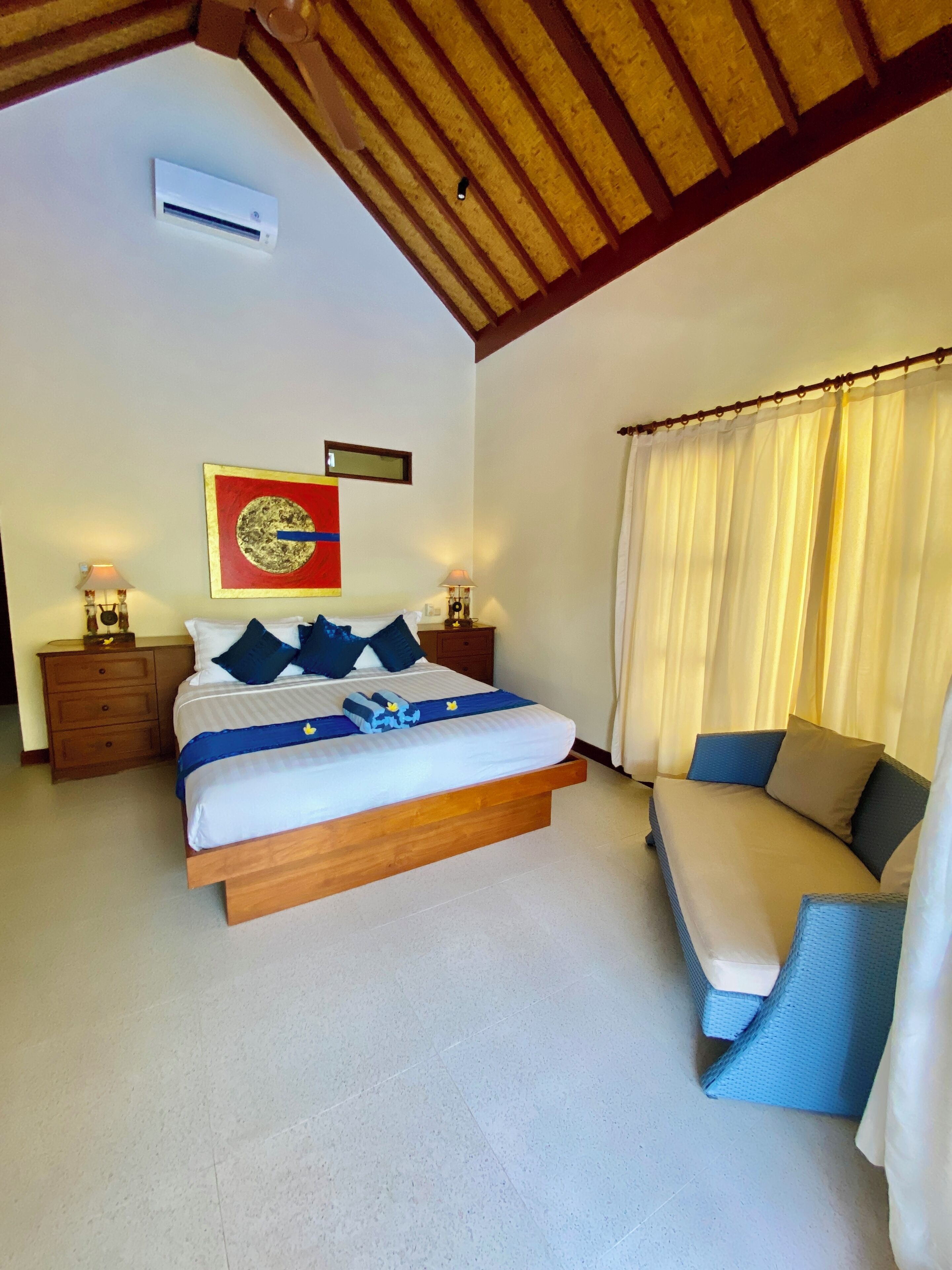Un dormitorio con cama, mesitas de noche, aire acondicionado instalado en la pared, una ventana con cortinas y un adorno en la pared.