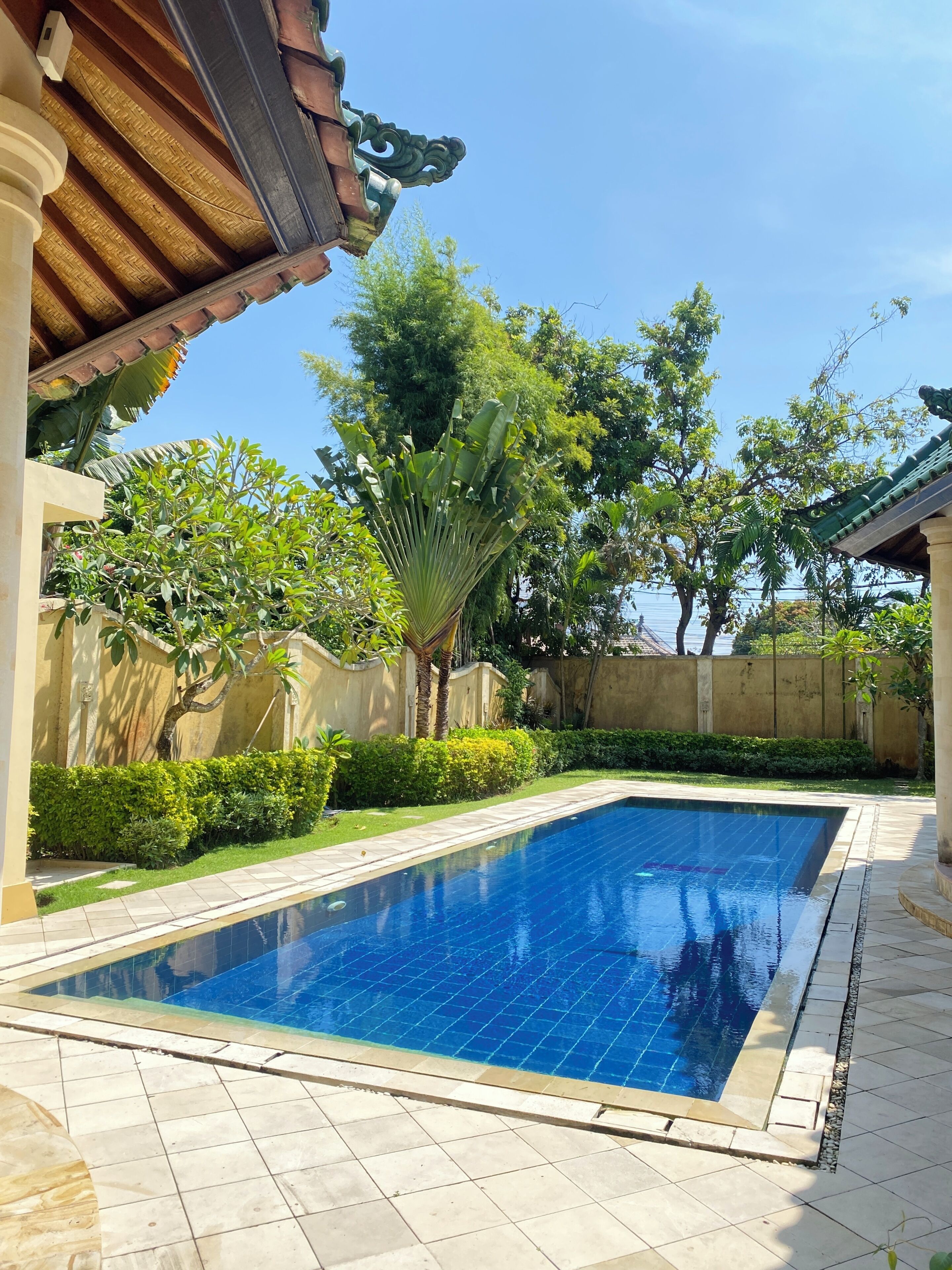 A swimming pool with tiled edges, surrounded by greenery and a building with a tiled roof.