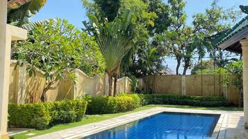 Una piscina con bordes de azulejos, rodeada de follaje y un edificio con techo de tejas.