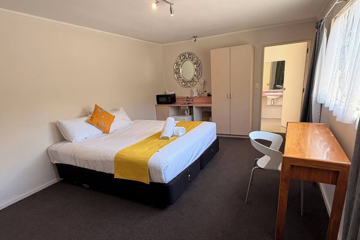 A hotel room with a bed, a desk, a mirror, a wardrobe, and a bathroom visible through an open door.