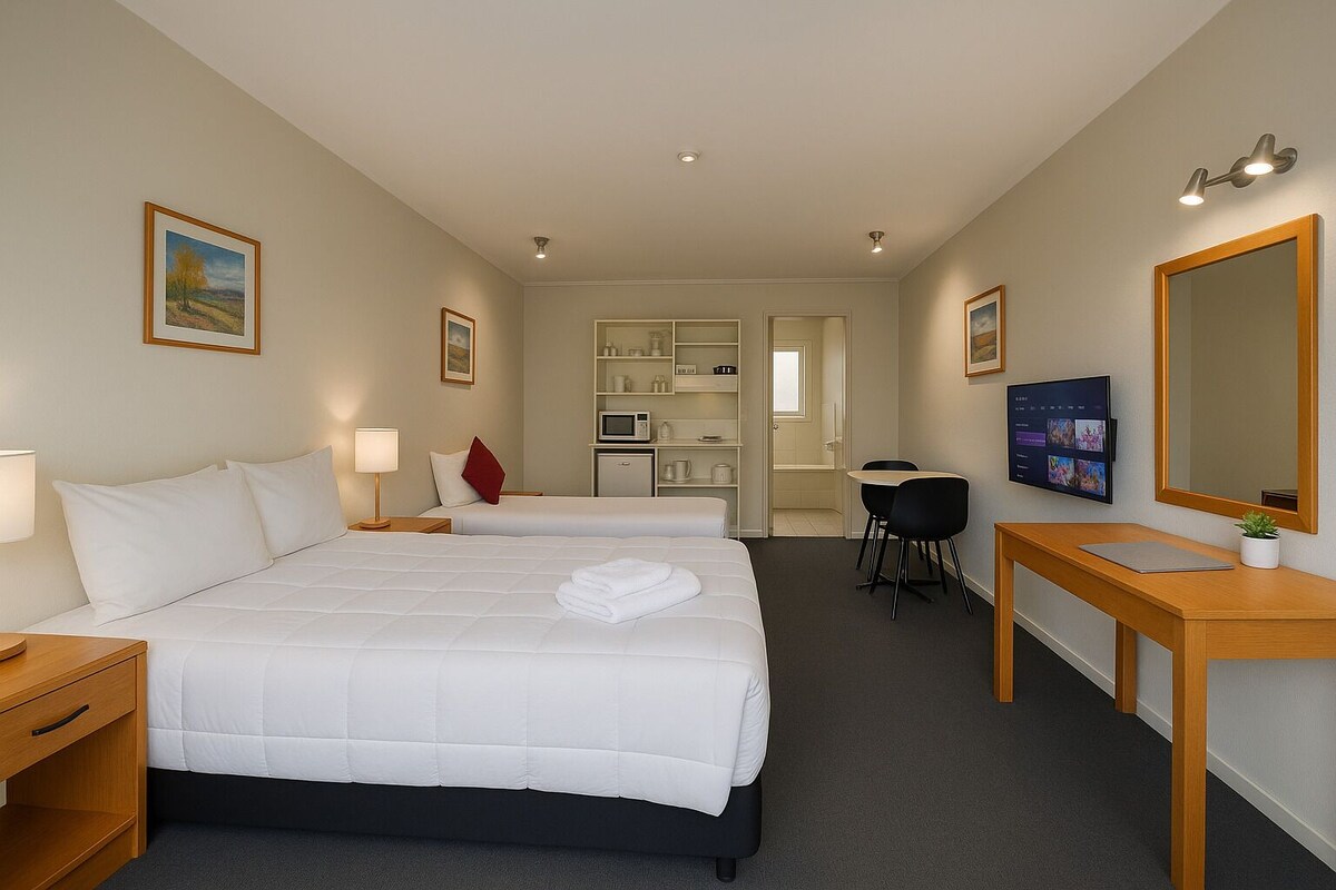 A hotel room with a bed, bedside tables, a desk, a chair, a TV, and a bathroom visible through an open door.