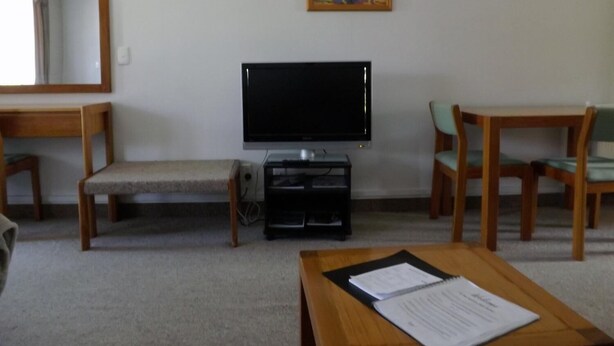 A living room with a television, two wooden chairs, a small table, and a painting on the wall. - Karaka Tree Motel (Taupo)