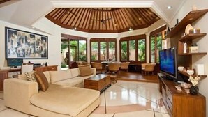 A spacious living room with a high ceiling, large windows, a television, and a wooden ceiling. - Bali Jade Villas (Denpasar)