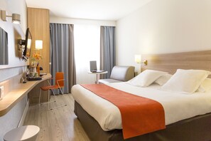 A hotel room with a bed, a desk, a chair, a small table, and a lamp.
