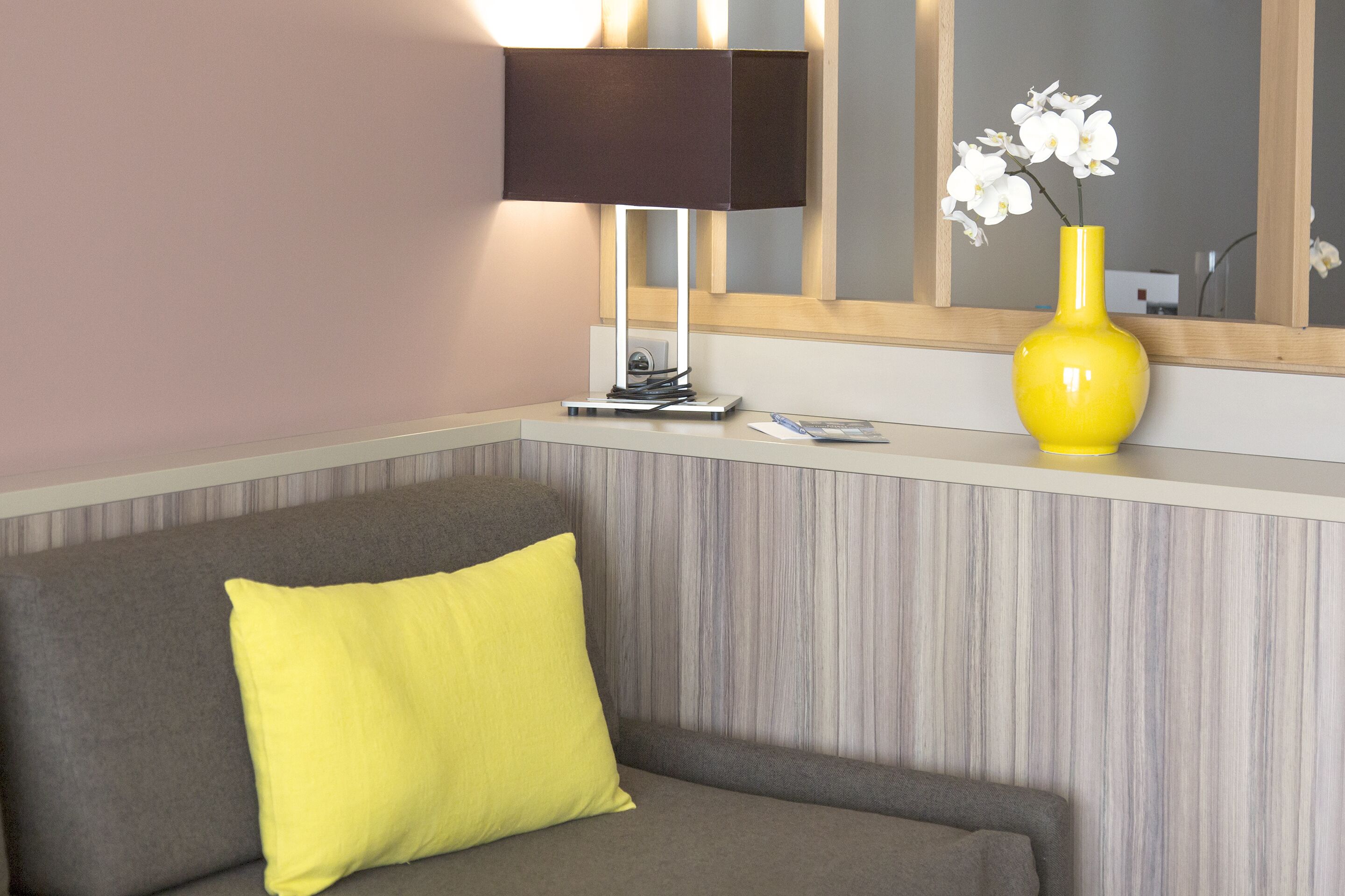 Un salon moderne avec un canapé gris, un coussin jaune, un vase jaune contenant des fleurs blanches, une lampe et un miroir au mur.