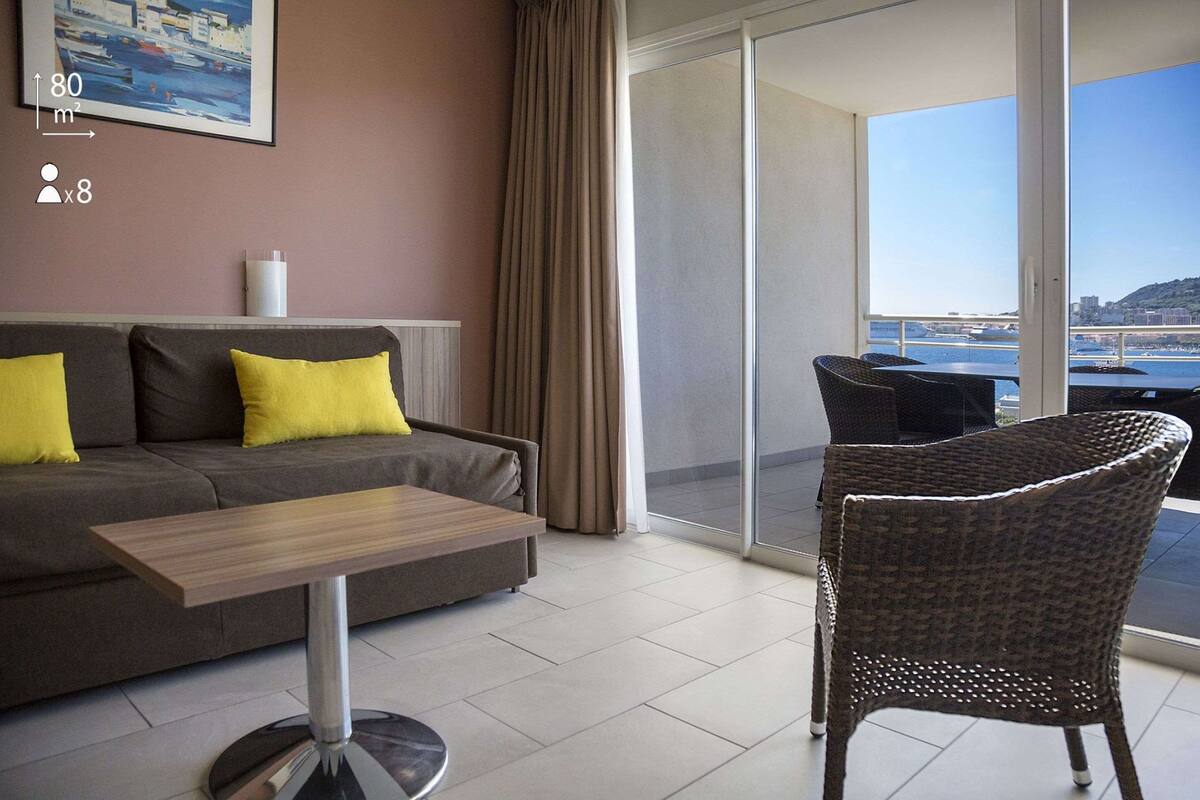 A modern living room with a sofa, a table, and a wicker chair. A large window offers a view of the sea and a bridge.