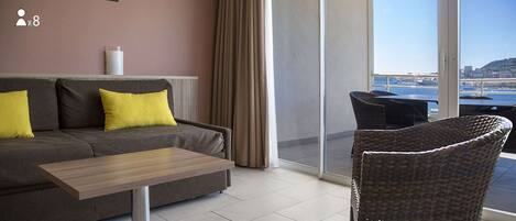 Un salón moderno con un sofá, una mesa y una silla de mimbre. Un ventanal grande ofrece vista al mar y un puente.