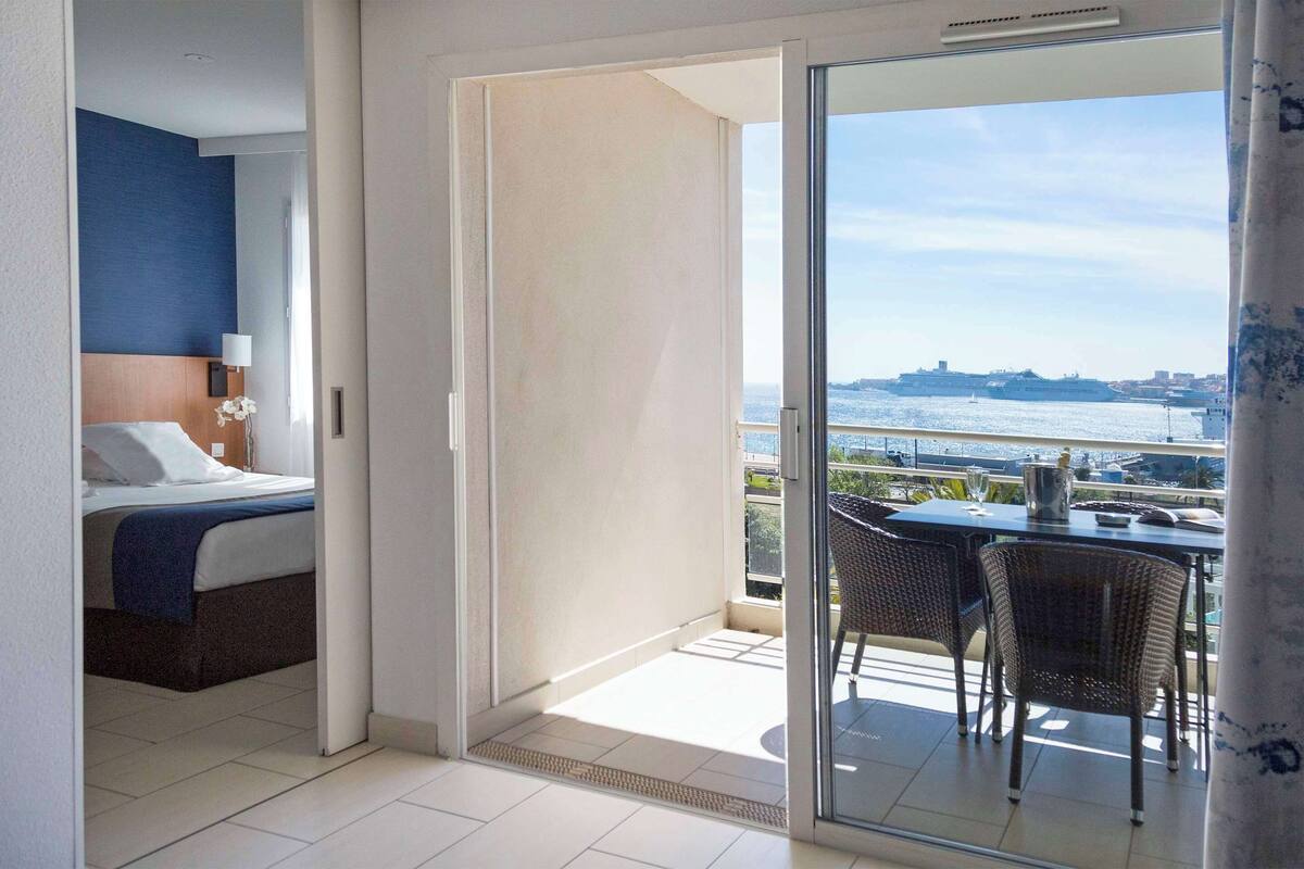 A hotel room with a bed, a small table with chairs, and a balcony with a view of the sea and a ship.