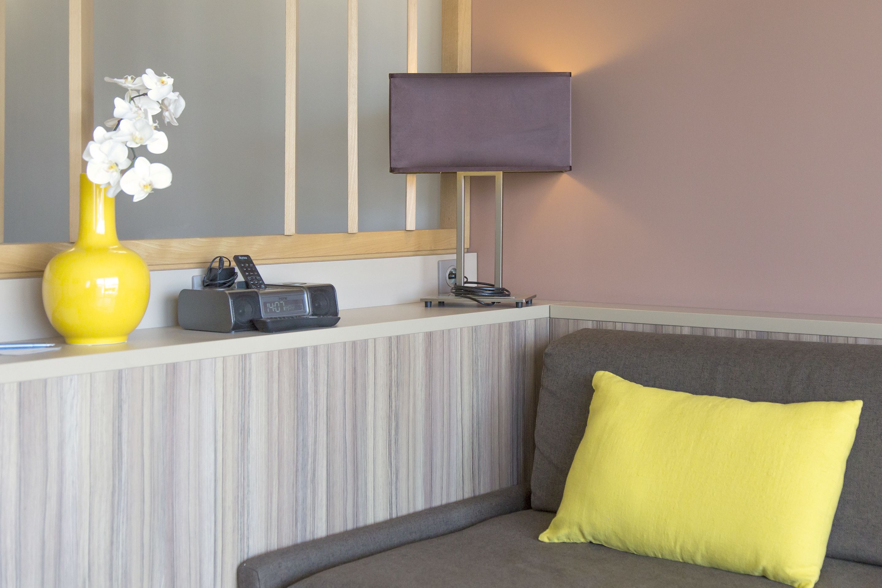 A room with a sofa, a yellow vase with white flowers, a lamp, and a small table with a telephone and a remote control.