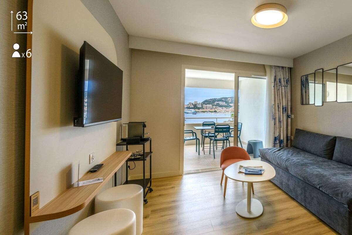 A modern living room with a sofa, a TV, a small table, and a balcony with a view of the sea.