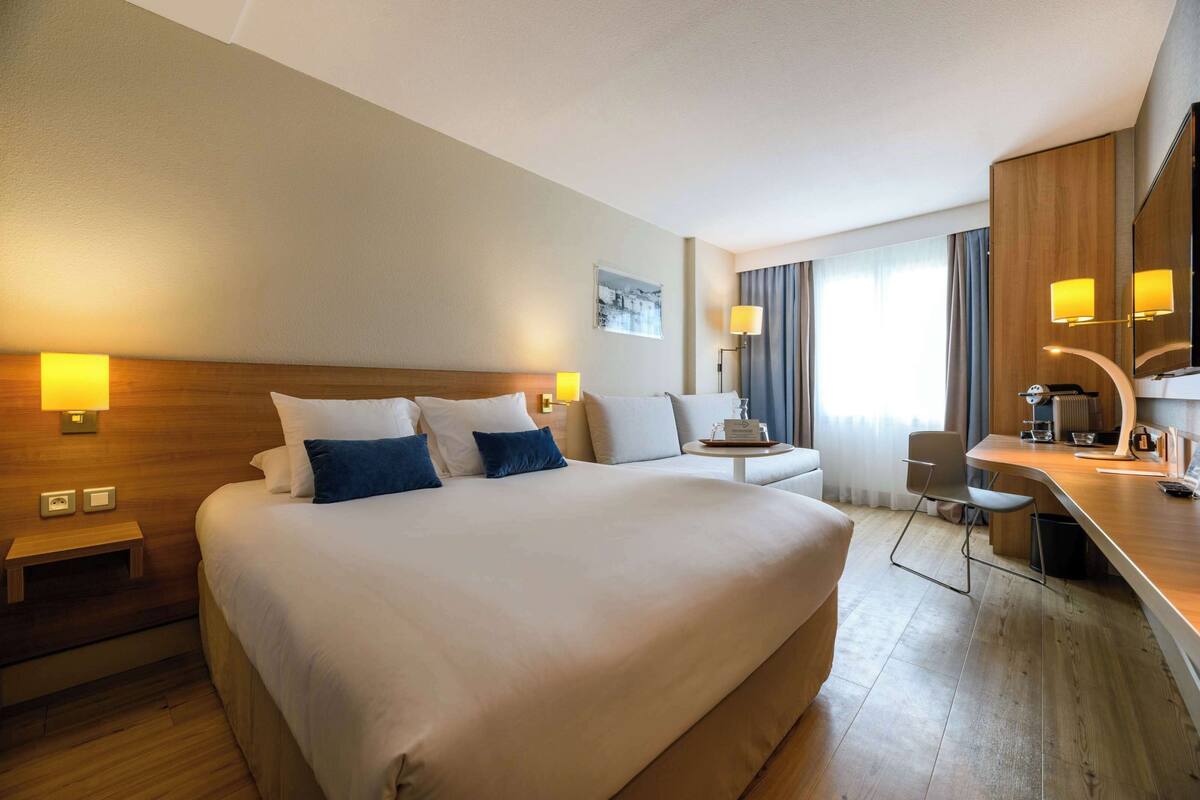 Ein Hotelzimmer mit einem großen Bett, Nachttischen, einem Schreibtisch mit Stuhl, einem Spiegel und einem Fenster mit Vorhängen.