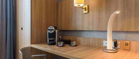 Una habitación de hotel moderna con un escritorio de madera, una cafetera, dos vasos, una silla y una lámpara de pared.