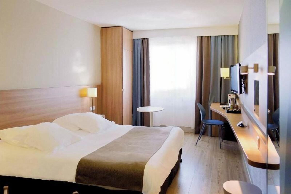 A hotel room with a bed, a desk, a chair, a TV, and a small table.