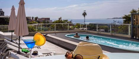 A poolside area with a clear view of the sea, a bar with a blue drink, and a straw hat on the table.
