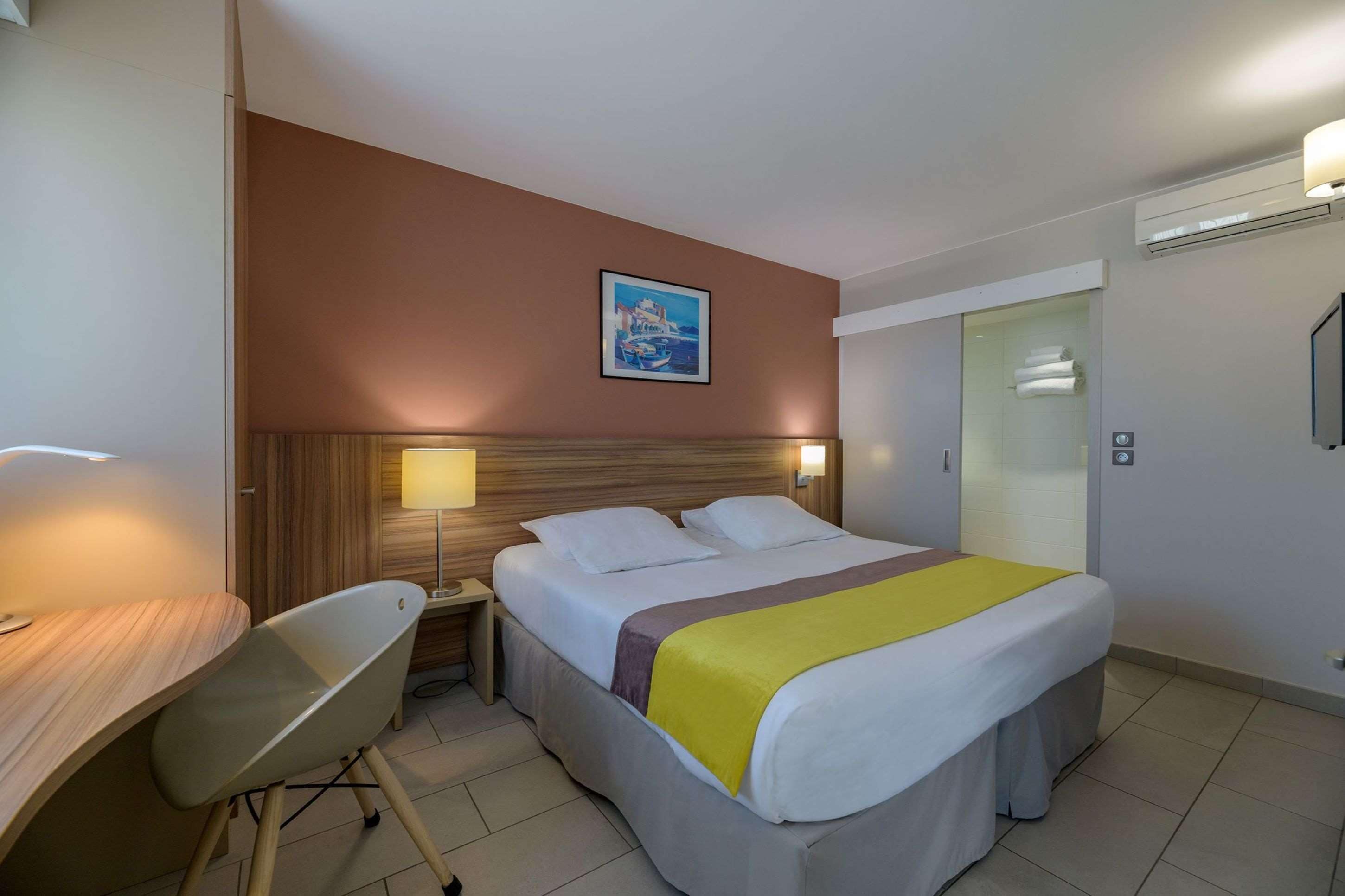 A hotel room with a bed, a desk, a chair, a nightstand, a lamp, a painting on the wall, and a sliding door.