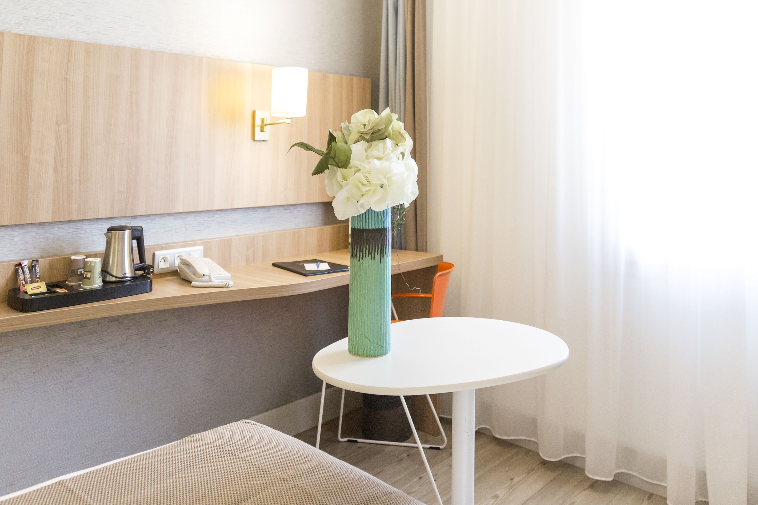 A hotel room with a wooden wall, a white round table, a green vase with white flowers, a coffee maker, a phone, and a trash bin.