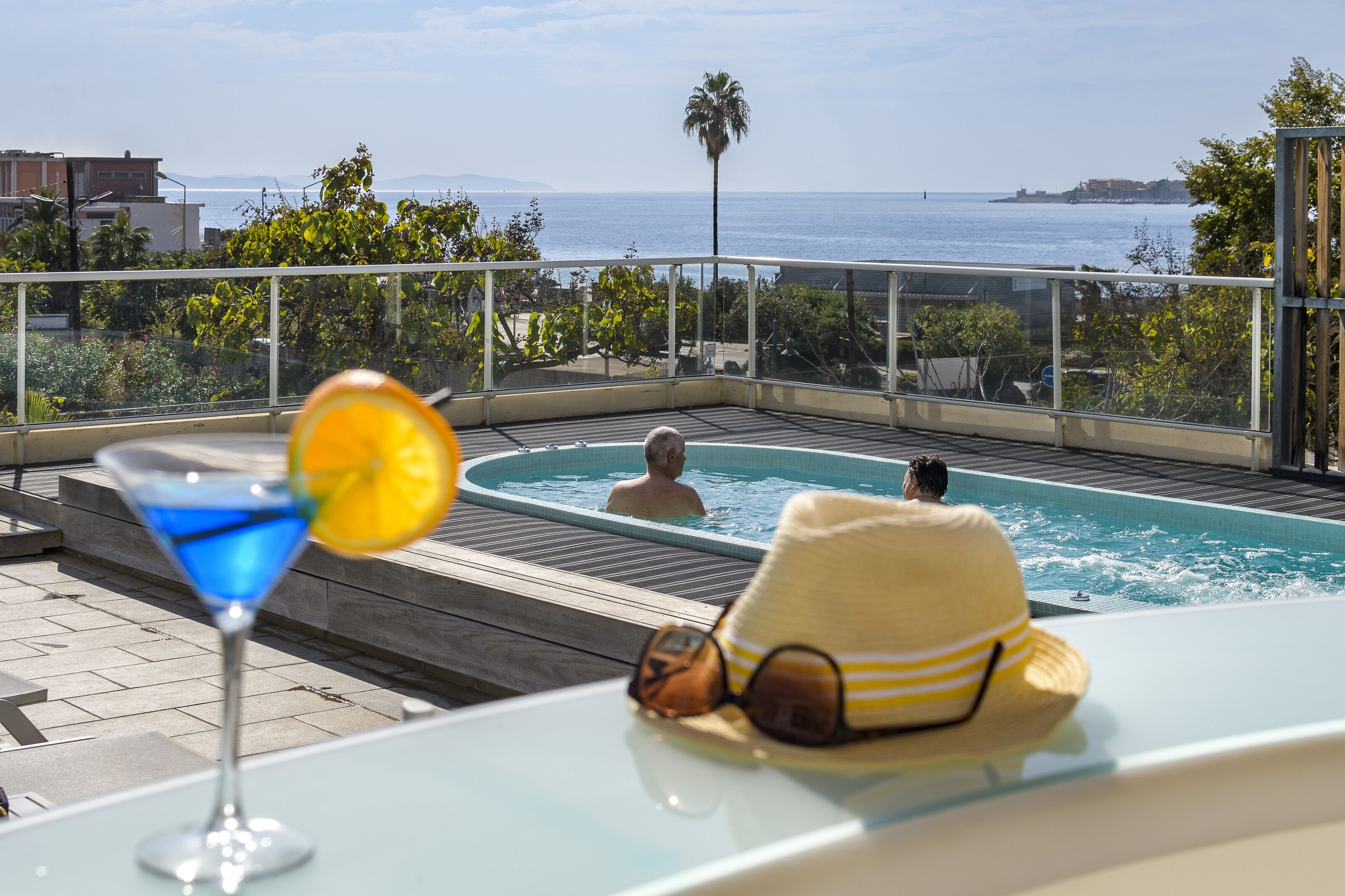 Ein Pool auf dem Dach mit freiem Blick auf das Meer, eine Lounge mit einem Getränk und einem Sonnenhut auf dem Tisch.