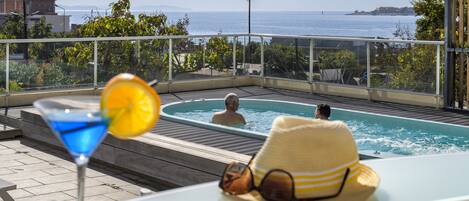 Ein Pool auf dem Dach mit freiem Blick auf das Meer, eine Lounge mit einem Getränk und einem Sonnenhut auf dem Tisch.