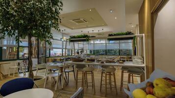 Un comedor moderno con mesas y sillas de madera, un gran árbol y vistas de la ciudad a través de amplios ventanales.